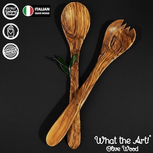 May include: A pair of salad servers crafted from Italian olive wood, featuring a spoon and a fork with distinct natural grain patterns. The set includes a small green olive leaf resting between the handles, showcasing the wood's organic texture.