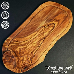 K&ouml;nnte beinhalten: Ein Olivenholz-Schneidebrett mit nat&uuml;rlichem Holzmaserungsmuster. Das Brett hat eine gefr&auml;ste Nut am Rand und den Text "What the Art! Olive Wood" unten rechts. Das Brett ist als 100% nat&uuml;rliches und einzigartiges Produkt gekennzeichnet.