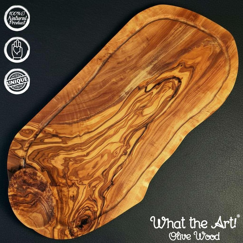K&ouml;nnte beinhalten: Ein Olivenholz-Schneidebrett mit nat&uuml;rlichem Holzmaserungsmuster. Das Brett hat eine gefr&auml;ste Nut am Rand und den Text "What the Art! Olive Wood" unten rechts. Das Brett ist als 100% nat&uuml;rliches und einzigartiges Produkt gekennzeichnet.