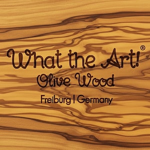 K&ouml;nnte beinhalten: Nahaufnahme einer Holzoberfl&auml;che mit dem Text "What the Art!" und "Olive Wood" in Dunkelbraun. Der Text "Freiburg | Germany" ist ebenfalls sichtbar. Das Holzmaserungsmuster ist sichtbar.
