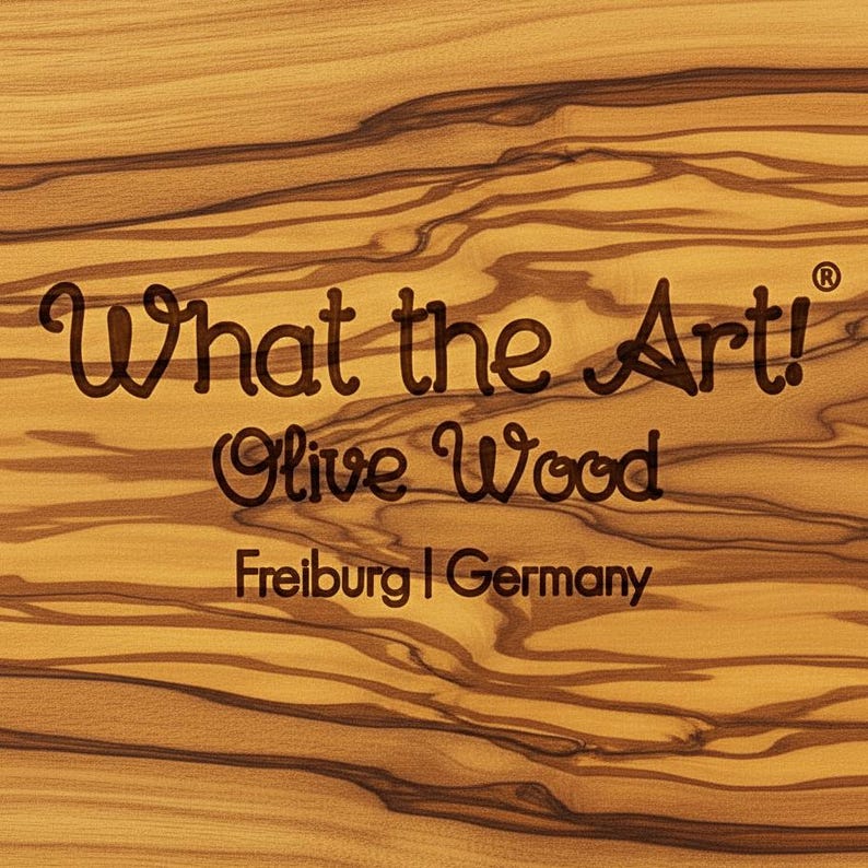 K&ouml;nnte beinhalten: Nahaufnahme einer Holzoberfl&auml;che mit dem Text "What the Art!" und "Olive Wood" in Dunkelbraun. Der Text "Freiburg | Germany" ist ebenfalls sichtbar. Das Holzmaserungsmuster ist sichtbar.