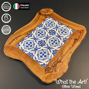 Op de afbeelding: Een olijfhouten serveerplank met een blauw en wit tegelontwerp. De plank heeft een unieke, onregelmatige vorm en een kleine, ronde uitsparing. De tekst "What the Art! Olive Wood" is zichtbaar.