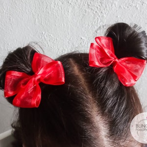 May include: Two red sheer ribbon bows are tied in the hair of a person with dark brown hair. The bows are tied in a bun on each side of the head.