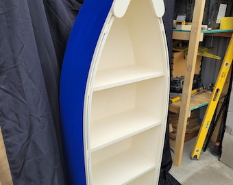 May include: A boat-shaped bookshelf, painted blue on the exterior and off-white inside. It features four shelves and a white top section. The design is reminiscent of a boat, offering a unique storage solution for any room.