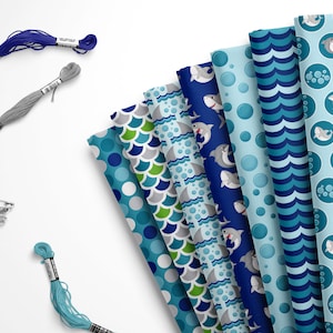 May include: A set of eight fabric squares with different blue and green shark-themed patterns. The patterns include sharks, bubbles, waves, and scales. The fabric squares are arranged in a fan shape and are surrounded by spools of blue, gray, and teal thread.