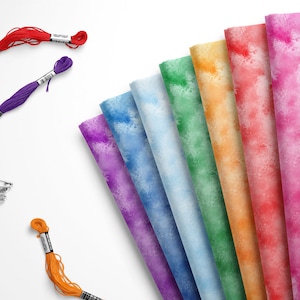 May include: A rainbow assortment of fabric swatches with a watercolor effect. The fabric is arranged in a fan shape with the colors transitioning from purple to pink.  There are also spools of embroidery floss in red, purple, and orange.