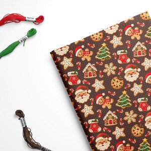 May include: A rectangular piece of fabric featuring a dark brown background with a repeating pattern of Santa Claus faces, gingerbread people, Christmas trees, stockings, candy canes, and cookies, accompanied by three skeins of embroidery floss.