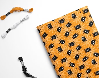 Op de afbeelding: Oranje stof met een herhalend patroon van zwarte spinnen en witte spinnenwebben. Er zijn drie spoelen borduurgaren op de afbeelding: oranje, wit en zwart.