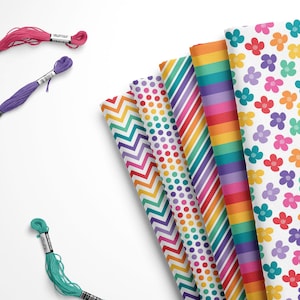 May include: A collection of colorful craft supplies, including spools of embroidery floss in pink, purple, and teal, alongside patterned paper rolls with floral, striped, and polka dot designs. The paper rolls feature rainbow colors.