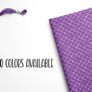 May include: Purple embroidery floss with 100 colors available. The floss is wound around a card and is shown next to a purple fabric with a mermaid scale pattern.