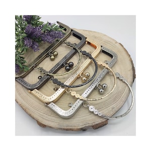 Può includere: Un assortimento di montature per borse in metallo con finiture varie, tra cui ottone antico, nero, oro e argento. Ogni montatura presenta dettagli decorativi e una chiusura a scatto. Le montature sono disposte su una superficie di legno, mostrando i loro diversi stili.