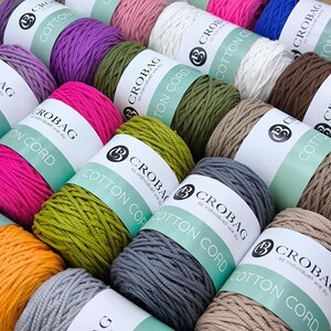 Könnte beinhalten: Eine Auswahl an farbigen Baumwollschnur-Spulen in verschiedenen Farbtönen, darunter Rosa, Grün, Lila und Grau. Jede Spule ist mit dem Markennamen "CROBAG" und "COTTON CORD" beschriftet. Das Bild zeigt eine Vielzahl von Bastelmaterialien.