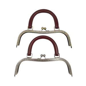 Op de afbeelding: Twee handtasgespen met rode handvatten. De bovenste gesp is van antiek messing met een decoratief ontwerp. De onderste gesp is van zilver met een decoratief ontwerp.