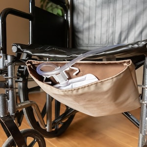 Könnte beinhalten: Ein schwarzer Rollstuhl mit einer beigefarbenen Tasche an der Seite. Die Tasche enthält einen durchsichtigen Plastikbeutel mit einem weißen Schlauch.