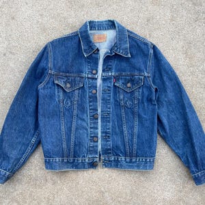 May include: A classic blue denim jacket with a button-up front, two chest pockets, and a small red tag. The jacket has a collared neckline and long sleeves. The denim is a medium wash, and the jacket appears to be in good condition.
