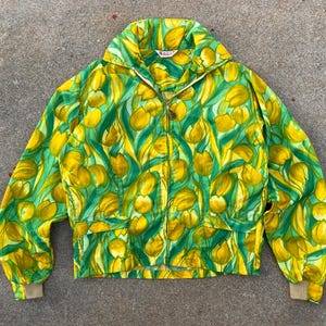 May include: A long-sleeve zip-up jacket featuring a vibrant all-over print of yellow tulips and green foliage. The jacket includes a high collar, a central metal zipper, two front pockets with flaps, and beige ribbed cuffs at the wrists.