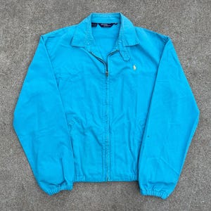 Puede incluir: Una chaqueta azul claro con cremallera y un pequeño logotipo bordado de jugador de polo en el pecho.