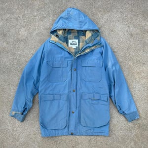 May include: A light blue hooded jacket with multiple front pockets and a plaid lining visible at the hood and collar. The jacket has a zipper closure and buttoned pockets. The Woolrich logo is visible on the inner lining.