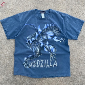 Può includere: T-shirt blu con una grafica dettagliata di Godzilla in sfumature di blu e bianco. La parola "GODZILLA" è stampata sotto in un carattere stilizzato. La maglietta ha un classico girocollo.