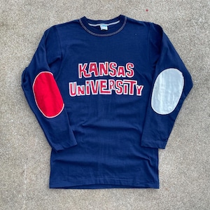 Könnte beinhalten: Marineblaues Langarmshirt mit dem Schriftzug "KANSAS UNIVERSITY" in roten und weißen Buchstaben. Das Shirt hat einen roten Ellbogenaufnäher am linken Ärmel und einen weißen am rechten.