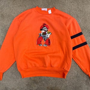 May include: Bright orange sweatshirt featuring a cartoon cowboy graphic on the front. The sleeves have two navy blue stripes. The sweatshirt has a crew neck and ribbed cuffs.