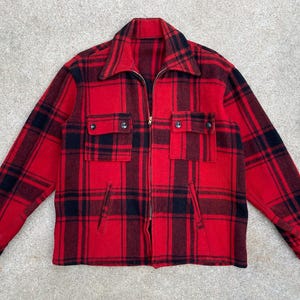 Puede incluir: Una camisa de franela a cuadros rojos y negros con cierre de cremallera. La camisa tiene dos bolsillos en el pecho y un solo bolsillo en el lado izquierdo.