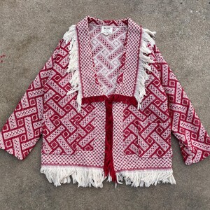 May include: A red and white patterned jacket with fringe trim. The jacket features a geometric design and a tie closure. The fringe is white, and the jacket has pockets. The tag reads "Big Jay".