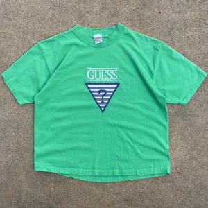May include: A green t-shirt with the "GUESS" logo in white, and a blue and white striped triangle design. The shirt is made of a soft material and has a classic crew neck.
