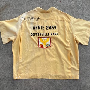 May include: A light yellow short-sleeved shirt with the name "McCullough" embroidered on the collar. The back of the shirt features the text "AERIE 2459 COFFEYVILLE, KANS." and a patch with a golden eagle.