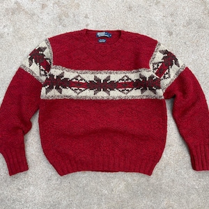 Puede incluir: Jersey de punto rojo con un patrón geométrico blanco y marrón alrededor del cuello. El jersey tiene cuello redondo y mangas largas.