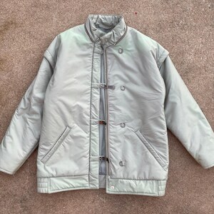 May include: A light green, quilted jacket with a high collar and a series of metal and leather closures down the front. The jacket has two front pockets and a ribbed hem. The jacket is a light, neutral color.