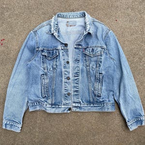 Könnte beinhalten: Eine hellblaue Jeansjacke mit klassischem Kragen, langen Ärmeln und einer Knopfleiste auf der Vorderseite. Das Kleidungsstück verfügt über zwei Brusttaschen mit geknöpften Klappen, zwei Seitentaschen und ein kleines rotes Etikett an der linken Brusttasche.