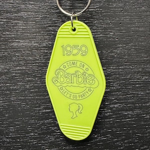 Könnte beinhalten: Ein neongrüner Schlüsselanhänger im Motel-Stil mit einem silbernen Schlüsselring. Der Schlüsselanhänger zeigt den Text "1959", "Come On Barbie, Let's Go Party" und eine Silhouette eines Frauenkopfes mit Pferdeschwanz. Ein Accessoire im Retro-Stil.