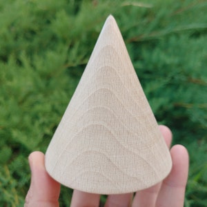 May include: A light wood cone shaped object with a textured surface. The cone is being held by a hand.