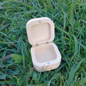 May include: An open, square wooden box with rounded corners, resting on green grass. The box is light-colored, possibly unfinished, with a small, round, light blue accent on the front and a gold-colored hinge. The lid is open, revealing the box's interior.