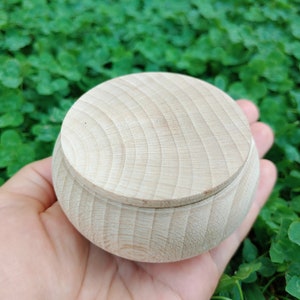 May include: A small, round, wooden box with a lid. The box is made of light-colored wood and has a smooth, polished finish.