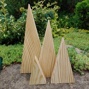 May include: Four wooden, triangular decorative trees of varying sizes. The trees are made of light-coloured wood with visible grain patterns. They are arranged outdoors on a stone surface, with greenery in the background.