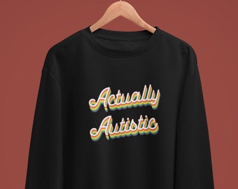 Może przedstawiać: Czarna bluza z okrągłym dekoltem i napisem Actually Autistic wydrukowanym czcionką w stylu retro z warstwowymi obrysami w kolorach żółtym, pomarańczowym i zielonym. Odzież posiada długie rękawy z prążkowanymi mankietami i dołem na drewnianym wieszaku.