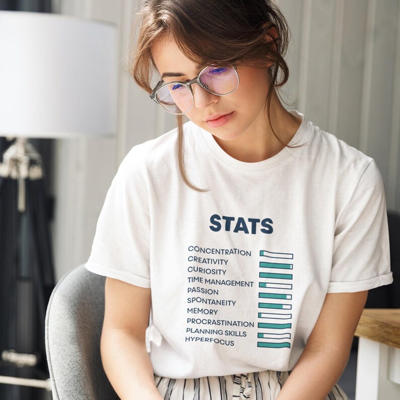 May include: A white short-sleeved t-shirt featuring the word STATS in dark blue text above a list of ten attributes including concentration, creativity, and curiosity, each paired with a horizontal progress bar graphic in teal and white.
