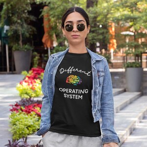 May include: A black t-shirt featuring the white text Different OPERATING SYSTEM and a colorful, abstract brain graphic. The shirt is worn under a light blue denim jacket. The design is centered on the chest, with the brain icon positioned between the lines.