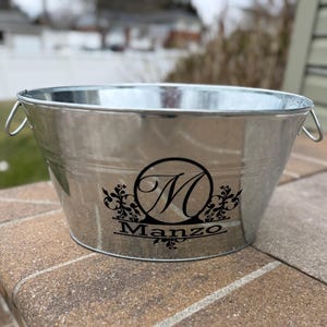 May include: A galvanized metal beverage tub with two handles. The tub features a black monogram with the letter "M" and the name "Manzo" in a decorative font. The tub is sitting on a brick surface.