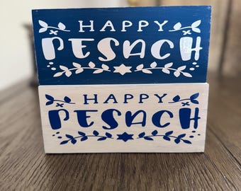 May include: Two wooden signs stacked on top of each other. The top sign is blue with white lettering that says "Happy Pesach". The bottom sign is white with blue lettering that says "Happy Pesach".