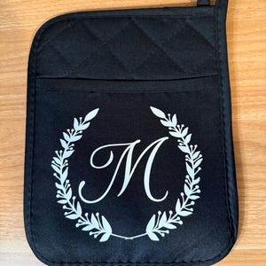 May include: Black quilted oven mitt with a white monogram "M" surrounded by a wreath of leaves.