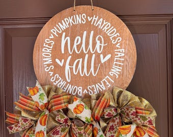 May include: A round wooden sign with the words "hello fall" in white script. The sign is decorated with a fall-themed ribbon and the words "pumpkins," "hayrides," "falling leaves," "bonfires," and "s'mores."