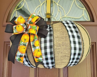 May include: A pumpkin-shaped wreath made of burlap, black and white checkered ribbon, and a decorative bow with pumpkin accents. The wreath is hanging on a brown door with a decorative window.
