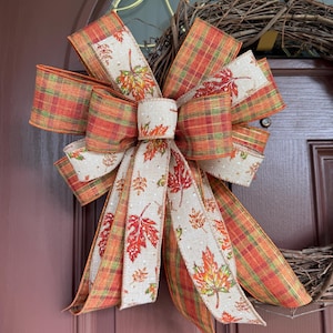 May include: A decorative fall bow with a plaid pattern in orange, red, and green. The bow features a central section with leaf designs in fall colors. The bow is attached to a grapevine wreath, perfect for autumn decor.