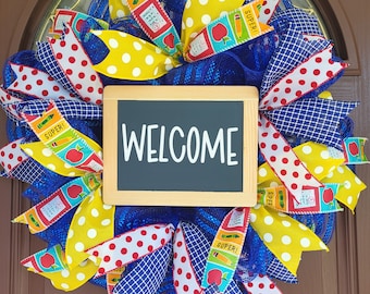 May include: A vibrant school-themed wreath with a blue mesh base. It features ribbons in yellow polka dots, red polka dots, and blue and white checkered patterns. A small chalkboard in the center reads "WELCOME".