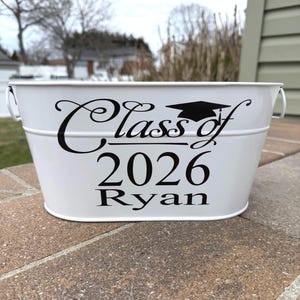 May include: A white metal bucket with black text that reads "Class of 2026 Ryan" and a graduation cap graphic. The bucket has two small handles and is sitting on a brick surface.