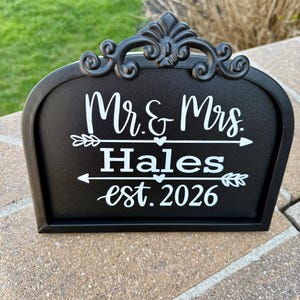 May include: A black sign with white text that reads "Mr. & Mrs. Hales est. 2026". The sign has a decorative scroll design at the top and arrows pointing towards the names. The sign is sitting on a brick surface.