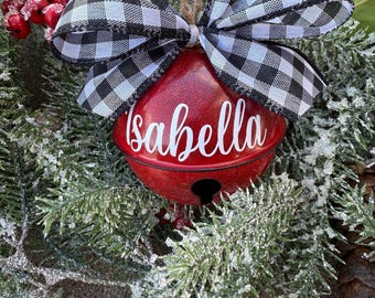May include: A red Christmas bell ornament with the name "Isabella" in white script. The bell is adorned with a black and white checkered bow and a jute string for hanging. The ornament is set against a backdrop of frosted evergreen branches and red berries.